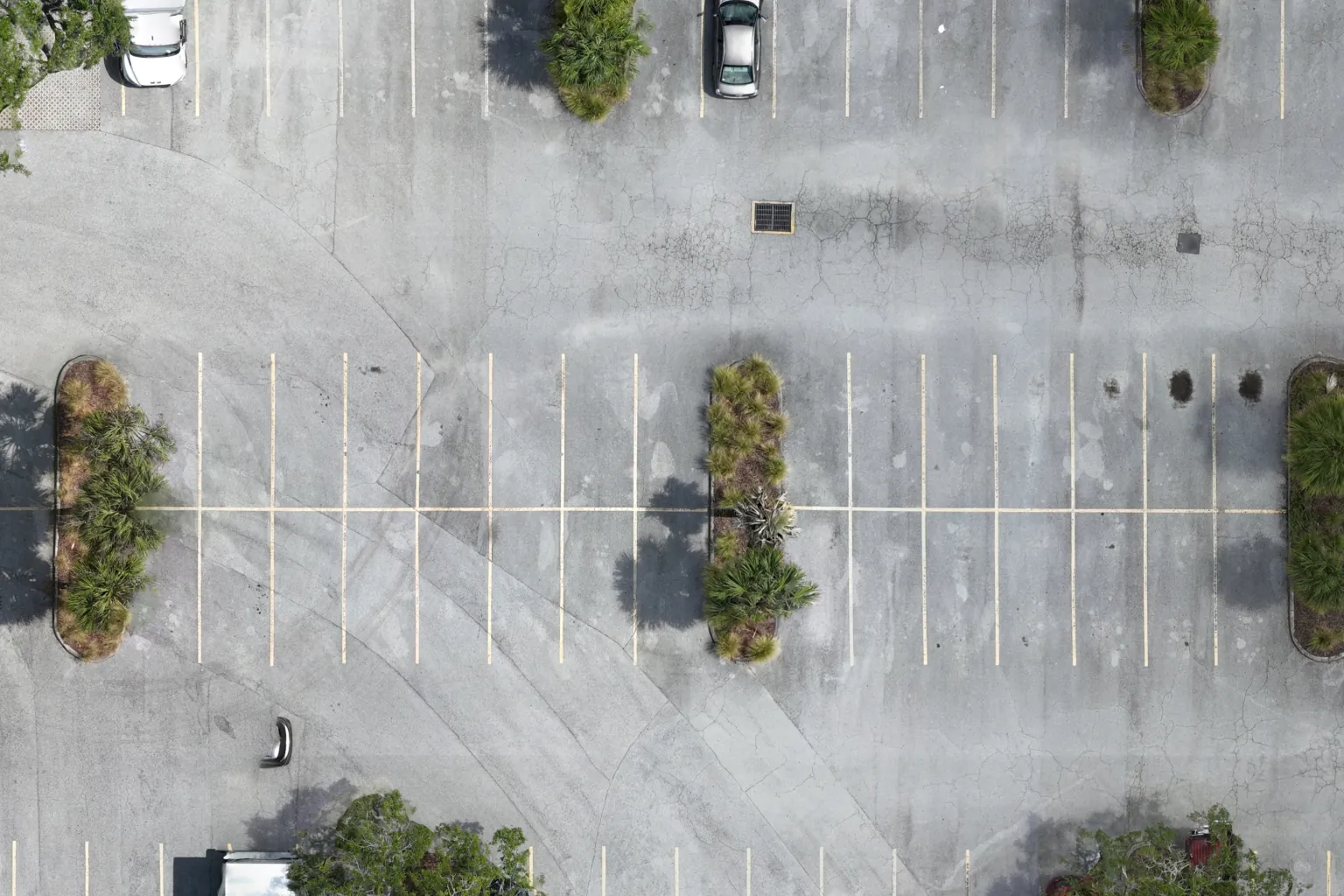 Native-resolution aerial detail of a parking lot: stall lines, a storm drain grate, alligator cracking in the asphalt, and a planted island.